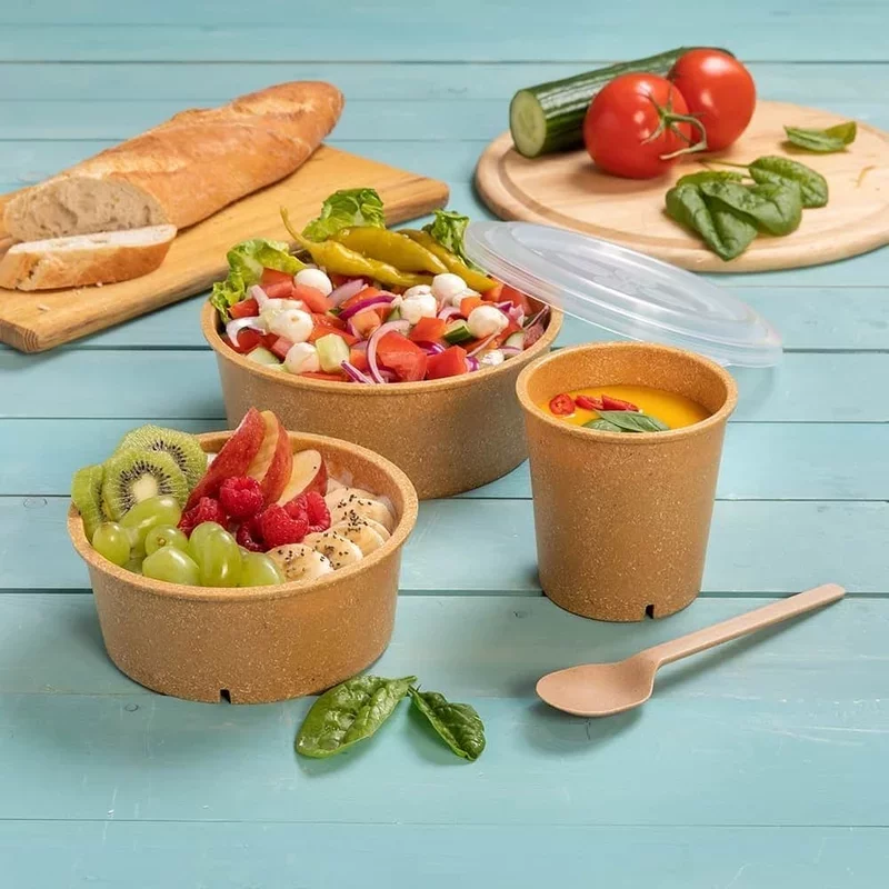 Bild eines angeschnittenen Baguettes auf einem rechteckigen Brettchen, zweier Salate, eines Bechers mit Löffel daneben und einer angeschnittenen Salatgurke mit zwie Tomaten auf einem runden Holzbrett